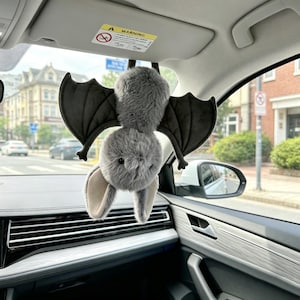 May include: A plush bat hanging upside down from a car visor. The bat has soft, light gray fur on its body and head, large pointed ears, and dark gray felt-like wings that are spread wide. It features small black eyes and a stitched nose detail.