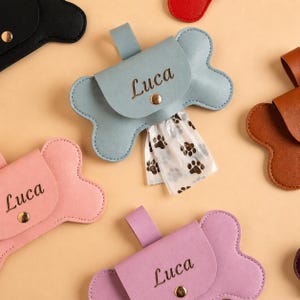 May include: A collection of bone-shaped dog waste bag dispensers made of smooth material in various colours including light blue, pink, purple, brown, and black. Each dispenser features a gold-tone snap button and the name Luca engraved on the front.