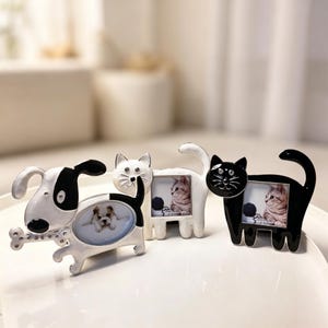 Puede incluir: Tres marcos de fotos decorativos con temática de mascotas. Un marco de perro en blanco y negro contiene una foto de un perro y un hueso. Dos marcos de gato, uno blanco y otro negro, cada uno contiene una foto de un gatito. Los marcos están sobre una superficie blanca.