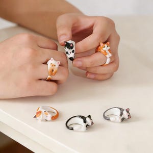 May include: A collection of cat-themed rings in various colors and poses. The rings feature detailed, miniature cat figurines in orange, black, white, and gray. Some rings are worn on fingers, while others are displayed on a white surface.