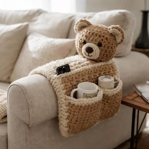 May include: A handmade, crochet teddy bear armrest organizer in shades of brown and cream. The bear head sits atop a blanket-style design with pockets for mugs and a phone. The armrest is placed on a beige sofa.