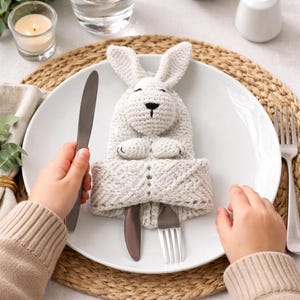 May include: A white crocheted bunny-shaped cutlery holder on a white plate. The holder has a black nose and eyes, and holds a knife and fork. The plate is set on a woven placemat, with a candle and napkin in the background.
