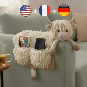 Puede incluir: Organizador de sofá con forma de vaca de las Tierras Altas en beige y marrón. Cuenta con dos bolsillos, uno con un libro y otro con un smartphone. La cabeza y los brazos de la vaca descansan sobre el brazo del sofá.