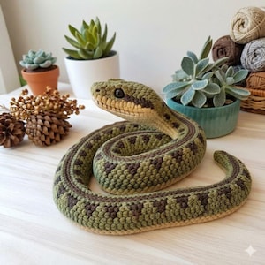 Könnte beinhalten: Gehäkelte Schlange in Grün- und Brauntönen, auf einer hellen Oberfläche aufgerollt. Die Schlange hat detaillierte Texturen und einen realistischen Kopf. Der Hintergrund umfasst Topfpflanzen und Garn, was auf einen handgefertigten Artikel hindeutet.