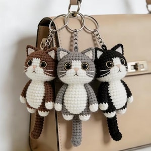 May include: Three crochet cat keychains in brown, gray, and black with white accents. Each cat has large, dark eyes and a small pink nose. The keychains are attached to silver metal rings, hanging in front of a beige handbag.