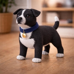 May include: A crocheted dog figurine in black and white, with a blue collar and a gold-colored tag. The dog has a white chest, paws, and a white stripe on its head. The tail has a white tip. The figurine is standing on a wooden floor.