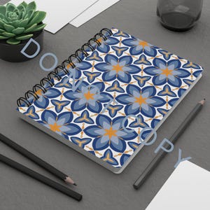 Puede incluir: Cuaderno de espiral con un patrón floral repetitivo en tonos azules, naranjas y blancos. El cuaderno está abierto, mostrando la cubierta estampada. También se ven dos lápices y una pequeña planta suculenta en una maceta negra.