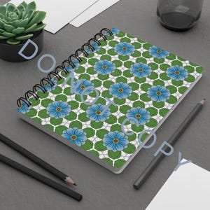 Puede incluir: Cuaderno con espiral con un patrón floral. La portada presenta un diseño repetitivo de flores azules con centros amarillos, sobre un fondo verde y blanco. El cuaderno está abierto, revelando páginas en blanco, y está acompañado de lápices.