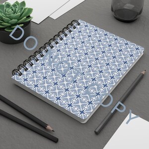 Puede incluir: Un cuaderno con espiral con un patrón geométrico azul y blanco. El cuaderno está abierto, revelando las páginas estampadas. Dos lápices negros y una pequeña planta suculenta en maceta también son visibles en la superficie gris.