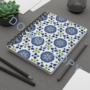 Puede incluir: Cuaderno de espiral con un patrón floral en tonos azules, verdes y blancos. El cuaderno está abierto, mostrando el diseño en sus páginas. También se ven dos lápices y una pequeña planta suculenta en maceta.