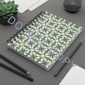 Puede incluir: Un cuaderno con espiral con una cubierta blanca con un patrón floral verde y gris. Dos lápices negros y una planta suculenta en una maceta negra también son visibles en una superficie gris.