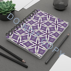Puede incluir: Un cuaderno de espiral con un patrón geométrico en morado, gris y blanco. El cuaderno está abierto, mostrando las páginas estampadas. También se ven dos lápices negros y una pequeña planta suculenta en una maceta negra.