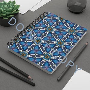 Puede incluir: Cuaderno con espiral con un patrón floral azul. El diseño presenta flores azules estilizadas con detalles dorados y morados sobre un fondo blanco. También se ven dos lápices y una pequeña planta suculenta.