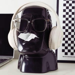 Puede incluir: Un dispensador de pañuelos con forma de estatua Moai negro brillante con gafas de montura transparente y auriculares blancos. Un pañuelo blanco sale de la nariz. El fondo incluye un portátil y un patrón de cuadros blancos y negros.