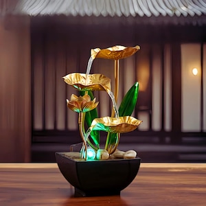 May include: An indoor water feature with a lotus flower design. The fountain has gold-coloured lotus leaves and a black square base. Water flows from the top leaves down to the base, with green and white accent lights.
