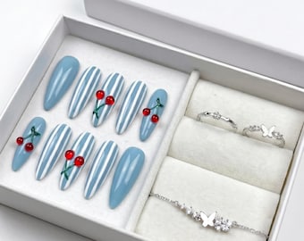 Può includere: Una scatola regalo bianca contenente dieci unghie finte a mandorla con strisce verticali blu e bianche, alcune con piccoli ciondoli a forma di ciliegia rossa. La scatola include anche due anelli argentati e un braccialetto argentato con farfalla.