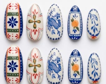 Può includere: Un set di dieci unghie finte a forma di mandorla con vari motivi dipinti a mano in blu, arancione, verde e rosso su base color crema. I disegni includono stelle geometriche, elementi floreali, un piccolo uccellino e frutti di arancia.