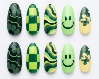 Può includere: Un set di dieci unghie finte a forma di mandorla con vari motivi verdi e gialli. I disegni includono fiori verde scuro, un motivo a scacchiera verde lime e bosco, strisce verdi ondulate, una faccina sorridente verde chiaro e un fiore giallo.