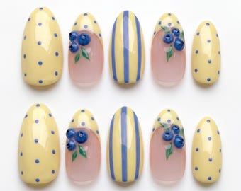 Può includere: Un set di dieci unghie finte a forma di mandorla con base giallo chiaro a pois blu, strisce verticali blu e dettagli tridimensionali di mirtilli blu con foglie verdi su una base rosa traslucida per una manicure decorativa.