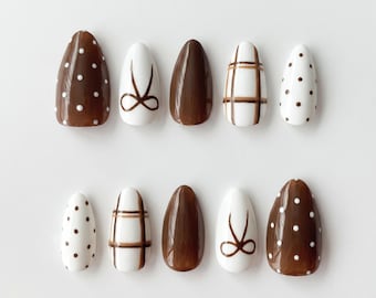 Può includere: Un set di dieci unghie finte a forma di mandorla con una palette di colori marrone e bianco. I design includono marrone tinta unita, bianco con pois marroni, marrone con pois bianchi, bianco con motivi a quadri marroni e bianco con fiocchi marroni.
