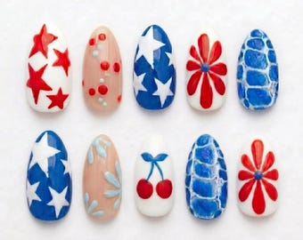 Può includere: Un set di dieci unghie finte a forma di mandorla con vari disegni, tra cui stelle rosse su bianco, puntini rossi su nude, stelle bianche su blu, fiori rossi su bianco, motivi blu testurizzati, stelle blu su bianco, foglie blu su nude e ciliegie.