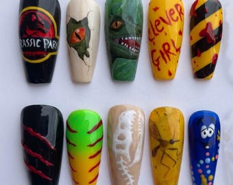Può includere: Un set di dieci unghie finte con vari disegni, tra cui il logo di Jurassic Park, un occhio di dinosauro, un volto di raptor, il testo 'clever GIRL', strisce di pericolo, graffi, uno scheletro, una zanzara e un personaggio blu.