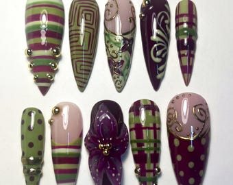 Può includere: Una collezione di dieci unghie finte a forma di mandorla. Le unghie presentano vari disegni, tra cui strisce, motivi geometrici, accenti floreali e pois. I colori includono tonalità di verde, viola, rosa e oro. Alcune unghie hanno abbellimenti dorati.