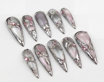 Può includere: Una collezione di unghie finte argentate e allungate. Ogni unghia presenta un design decorativo con accenti rosa e trasparenti, inclusi gemme. Le unghie sono disposte su una superficie bianca, mostrando i loro dettagli intricati.