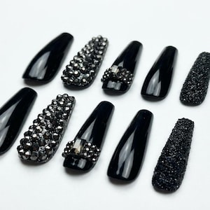 May include: A set of ten press-on nails in a coffin shape, featuring a glossy black finish. Some nails are adorned with clusters of black rhinestones, while others have a textured, granular black surface or a single central crystal accent.