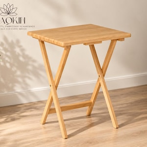 May include: A light-coloured wooden folding table with a rectangular top and an X-frame base. The table features a smooth, natural wood grain finish. The logo WAOKIN with a lotus icon and the text CREATIVE CRAFTS, EMBROIDERY & HANDMADE ESSENTIALS FOR EVERY MAKER is visible in the upper left corner.