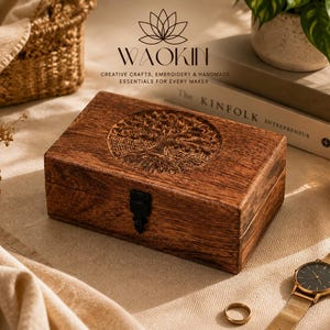May include: A rectangular wooden box with a dark brown finish and a visible wood grain. The lid features a circular carved design of a tree. A black metal latch is centred on the front. The text WAOKIN CREATIVE CRAFTS, EMBROIDERY & HANDMADE ESSENTIALS FOR EVERY MAKER is visible above the box.