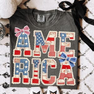 Op de afbeelding: Donkergrijs t-shirt met het woord "AMERICA" geappliqueerd in rood, wit en blauw stof. De letters zijn versierd met strikken en kanten rand. Het shirt wordt gecombineerd met witte laarzen.