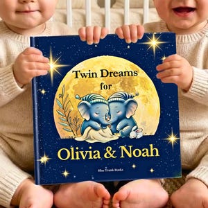 Peut inclure: Un livre relié intitulé Twin Dreams for Olivia & Noah avec une couverture bleu foncé illustrée d'une grande lune jaune, de deux éléphants bleus portant des bonnets de nuit rayés et d'étoiles dorées. Le titre et les noms sont visibles.