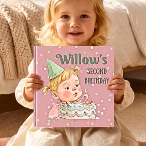 Può includere: Un libro quadrato con copertina rigida color rosa polvere, caratterizzato da un'illustrazione ad acquerello di un bambino con un cappellino da festa verde che spegne una candelina su una torta. Il testo recita Willow's SECOND BIRTHDAY in verde.
