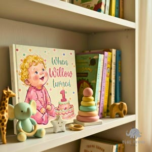 K&ouml;nnte beinhalten: Ein Kinderbuch mit dem Titel When Willow turned 1, das ein Baby im rosa Schlafanzug neben einer Geburtstagstorte mit einer Kerze in Form der Zahl Eins zeigt. Das Buch steht in einem Regal neben einer Giraffe, einem Elefanten und Stapelringen.