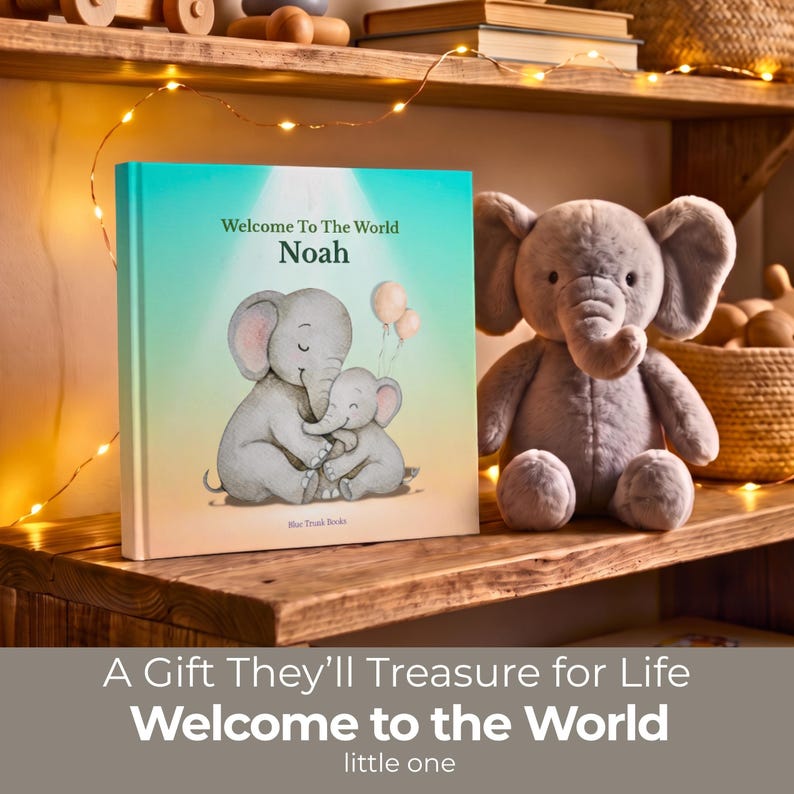 K&ouml;nnte beinhalten: Ein quadratisches Hardcover-Buch mit dem Titel Welcome To The World Noah mit einem hellblau-gr&uuml;nen Farbverlauf auf dem Einband, das zwei graue Elefanten und zwei Luftballons zeigt. Neben dem Buch steht ein grauer Pl&uuml;schelefant auf einem Holzregal.