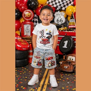Può includere: Un bambino indossa una maglietta bianca e pantaloncini di jeans, entrambi decorati con grafiche di auto da corsa. La maglietta presenta un numero 1 a scacchi e il nome "Brandon". Lo sfondo include palloncini, un cartello della Route 66 e uno striscione di compleanno.