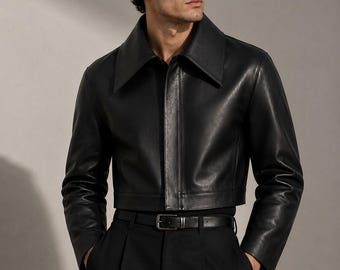 May include: A black cropped leather jacket featuring a wide pointed collar, long sleeves, and a concealed front closure. The jacket is paired with black high-waisted trousers and a black leather belt with a simple rectangular metal buckle.