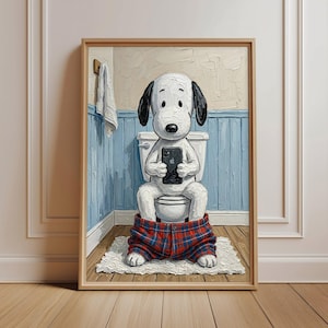 Puede incluir: Una obra de arte enmarcada representa a un perro de dibujos animados sentado en un inodoro, absorto en un teléfono inteligente. El perro lleva pantalones de cuadros rojos y azules. El baño presenta paneles azules, una toalla blanca y una alfombra blanca.