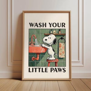 Op de afbeelding: Een ingelijste print met Snoopy die zijn poten wast in een rode wastafel. De tekst "WASH YOUR LITTLE PAWS" staat in het zwart. De achtergrond is een groene tegelmuur met een plank en een handdoekenrek.