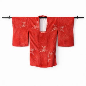 May include: A red kimono with a floral pattern. The garment is displayed on a black rod against a white background. The kimono has wide sleeves and a square neckline. The floral design is in a lighter shade of red.
