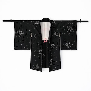 May include: Black kimono with white polka dot pattern, displayed on a black rod. The kimono has a white inner lining with pink accents and a pink tassel. The sleeves are long and the garment is open in the front.