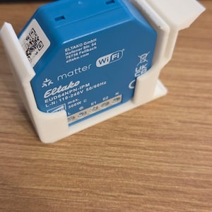 Könnte beinhalten: Ein blaues Eltako EUD64NPN-IPM Wi-Fi Smart-Home-Gerät in einem weißen 3D-gedruckten Halter. Das Gerät trägt die Aufschrift "matter" und "Wi-Fi". Das Gerät befindet sich auf einer Holzoberfläche.