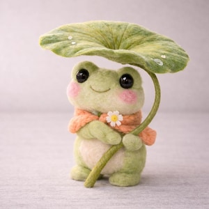 Könnte beinhalten: Handgefertigte Froschfigur aus Filz, die ein grünes Blatt über sich hält. Der Frosch hat rosige Wangen, große schwarze Augen und trägt einen orangefarbenen Schal mit einer weißen Blume. Ein skurriles, dekoratives Objekt.