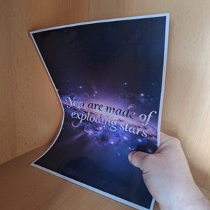Puede incluir: Una impresión con un diseño de nebulosa cósmica en tonos morados, azules y blancos. El texto "You are made of exploding stars" está escrito en la imagen. La impresión se sostiene en una estantería de madera.