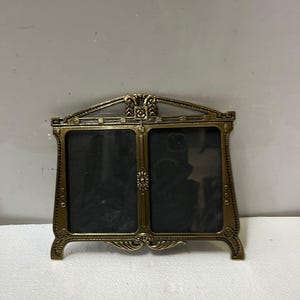 Puede incluir: Un marco de fotos doble de estilo vintage, con acabado en bronce. El marco tiene un diseño ornamentado con elementos decorativos en la parte superior y central. Está diseñado para contener dos fotos.