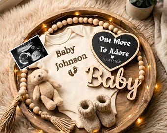 May include: A wooden tray with baby items, including a onesie with "Baby Johnson" printed on it, a teddy bear, knitted booties, and a heart-shaped chalkboard sign that says "One More To Adore". An ultrasound image is also present.
