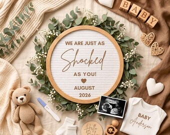 May include: A round felt letter board with a wooden frame displaying the text WE ARE JUST AS Shocked AS YOU! AUGUST 2026, surrounded by a green eucalyptus wreath, a teddy bear, a pregnancy test, baby socks, a rattle, and a baby onesie.