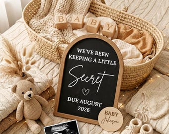 May include: An arched black chalkboard sign with white text reading WE'VE BEEN KEEPING A LITTLE Secret DUE AUGUST 2026, accompanied by a baby ultrasound photo, a wooden disc labeled BABY Johnson, a beige teddy bear, and a pair of knit baby booties.
