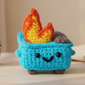 Op de afbeelding: Een gehaakte blauwe vuilnisbak met een lachend gezicht, zwarte ogen en grijze wielen. Oranje en gele vlammen komen uit de bovenkant. De vuilnisbak is een grillig, handgemaakt item.